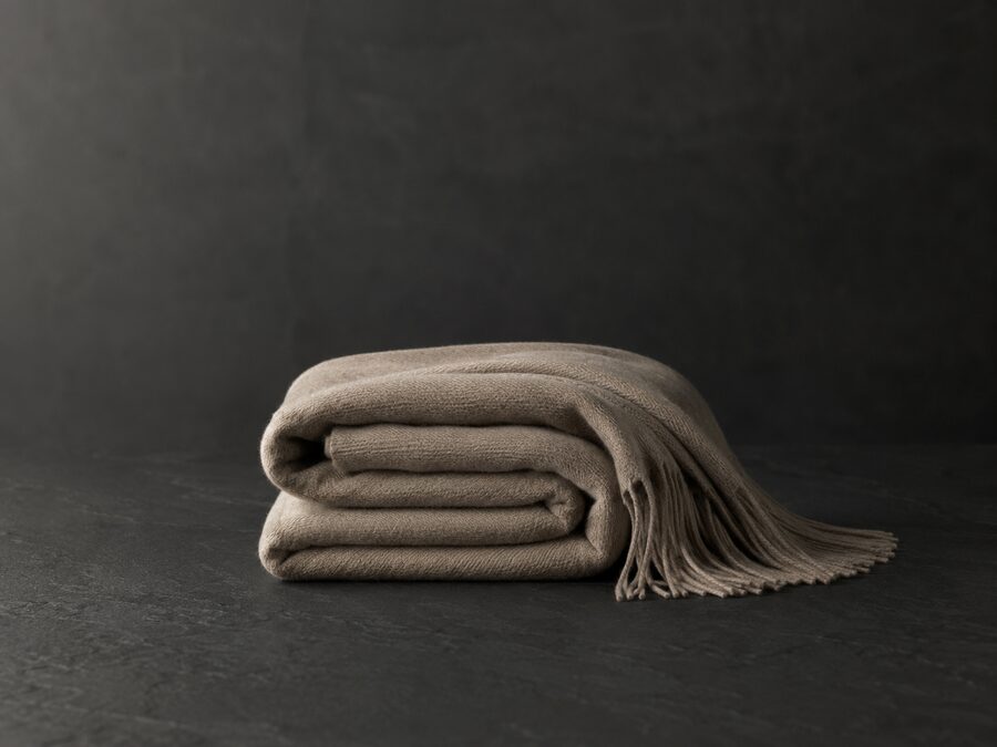 A folded cashmere throw with a fringed edge, on dark stone.